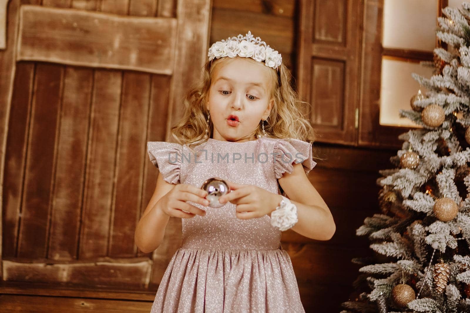 Little girl with blond hair in pink dress in new year background. Girl blue eyes near Christmas tree with ball in her hands.