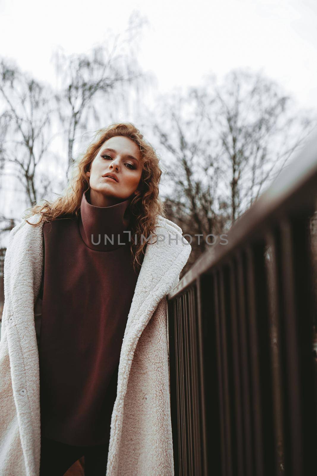 A girl with red curly hair in a white coat poses on outdoor parking in cold autumn. City Style - Urban