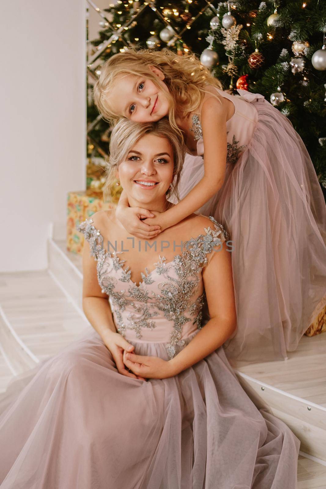 Daughter hugging her mother while sitting at christmas tree with gift boxes by natali_brill