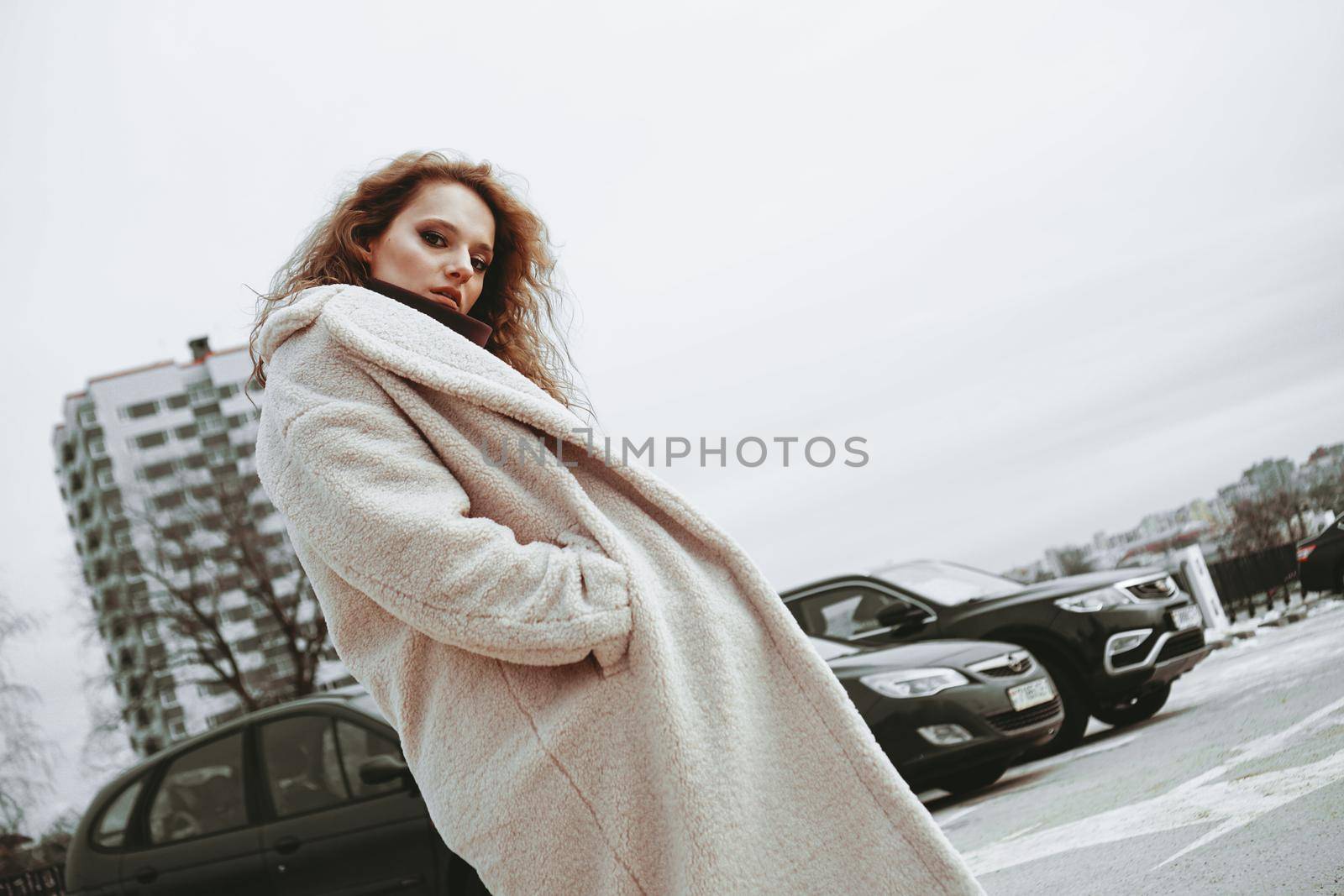 A girl with red curly hair in a white coat poses on outdoor parking in cold autumn. City Style - Urban