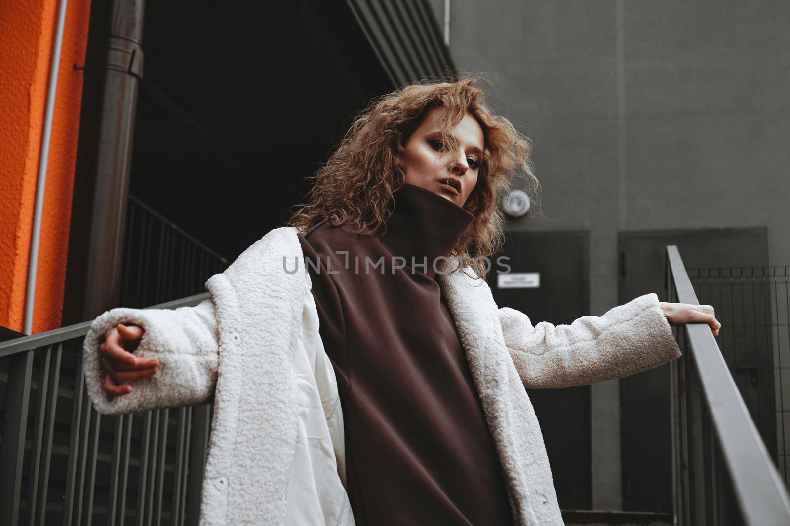 A girl with red curly hair in a white coat poses on the parking stairs by natali_brill