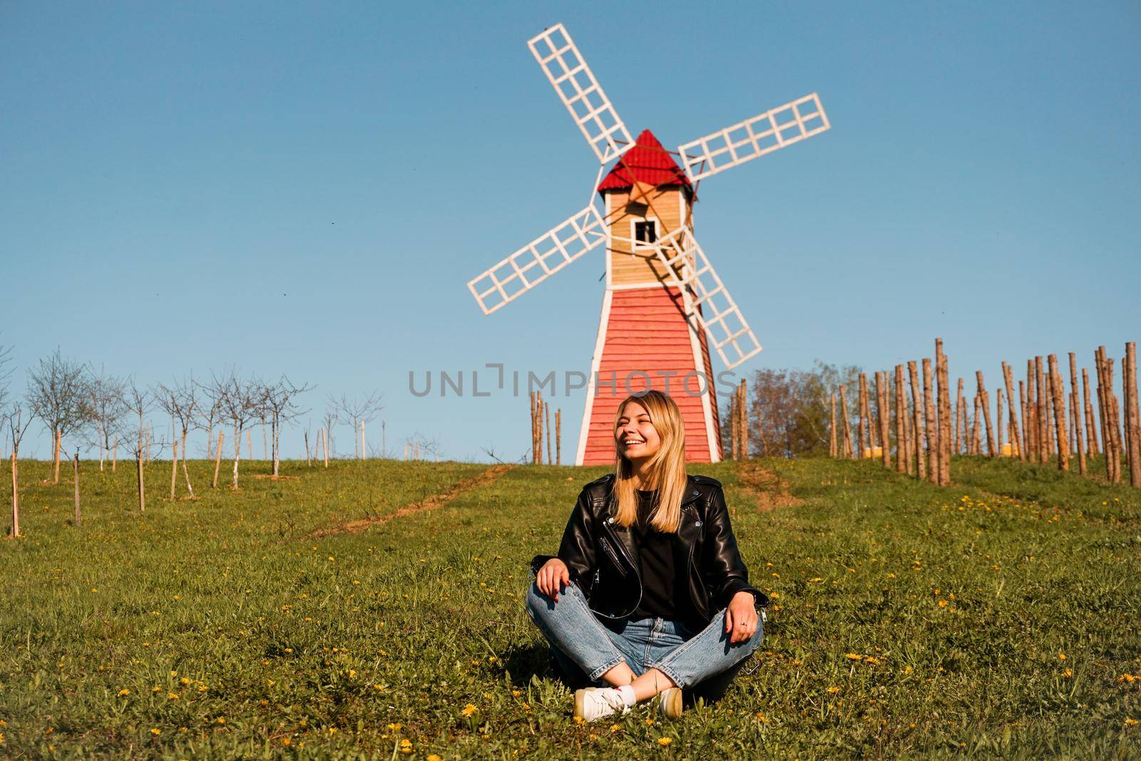 Beautiful young woman sits on the grass against the backdrop of the red mill. Sunny summer day. Rest in the countryside