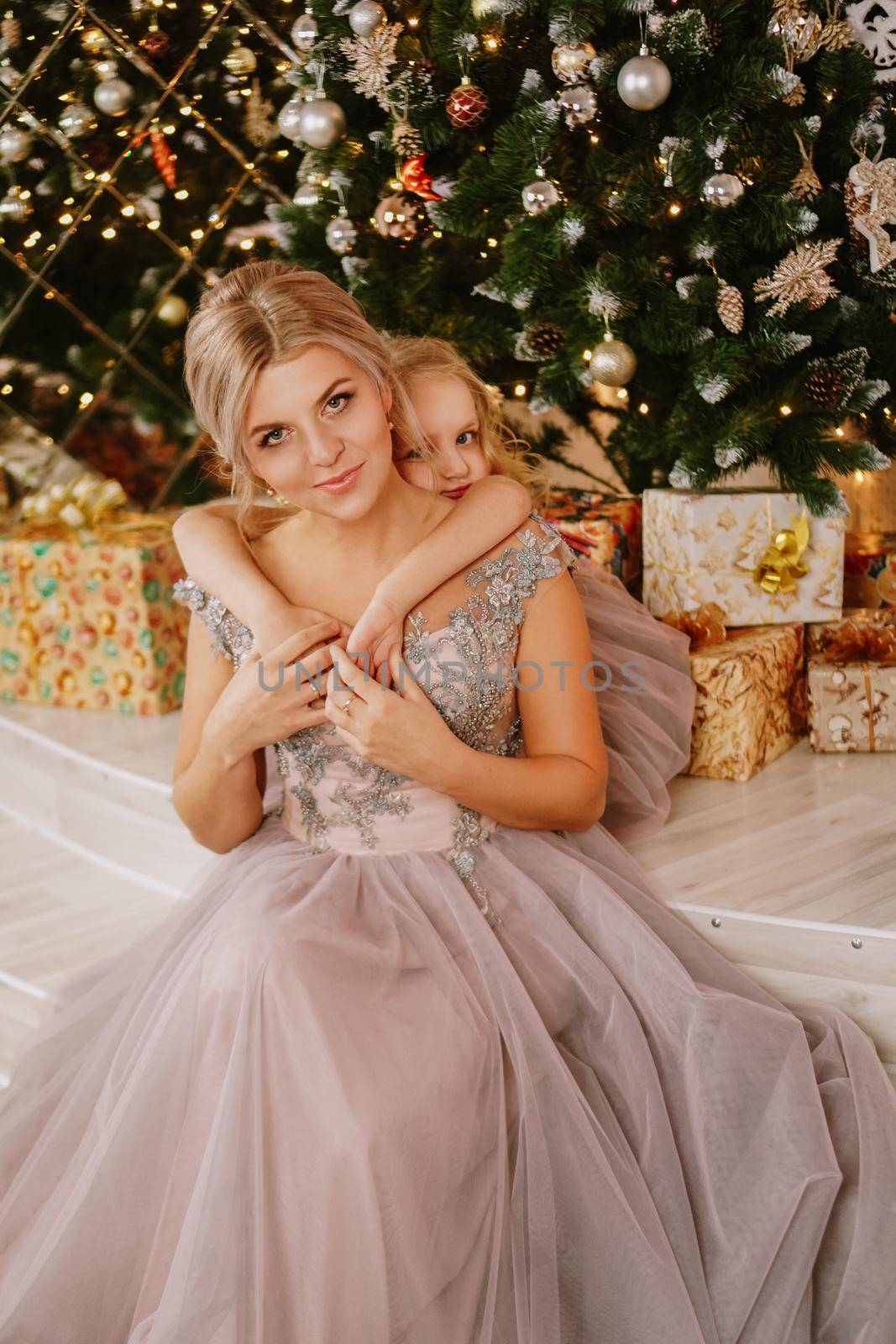 Smiling daughter hugging her mother while sitting at christmas tree with gift boxes