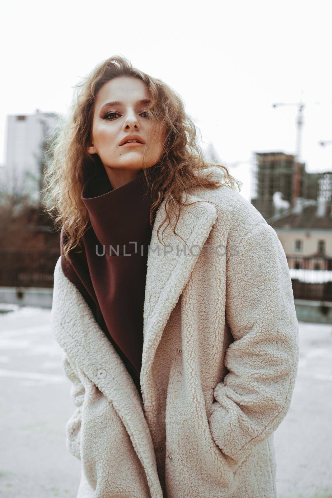 A girl with red curly hair in a white coat poses on outdoor parking in cold autumn. City Style - Urban. vertical photo
