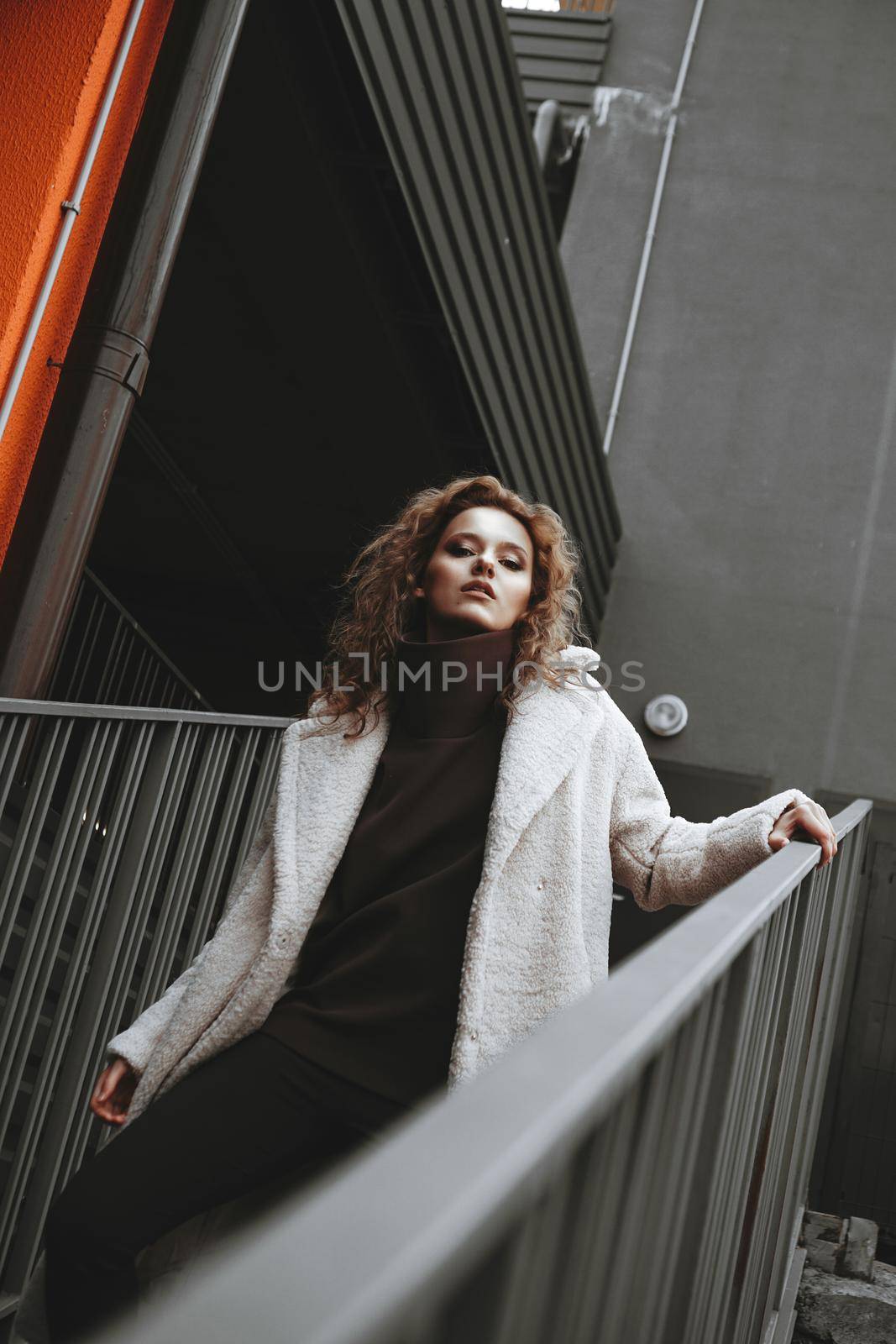 A girl with red curly hair in a white coat poses on the parking stairs by natali_brill