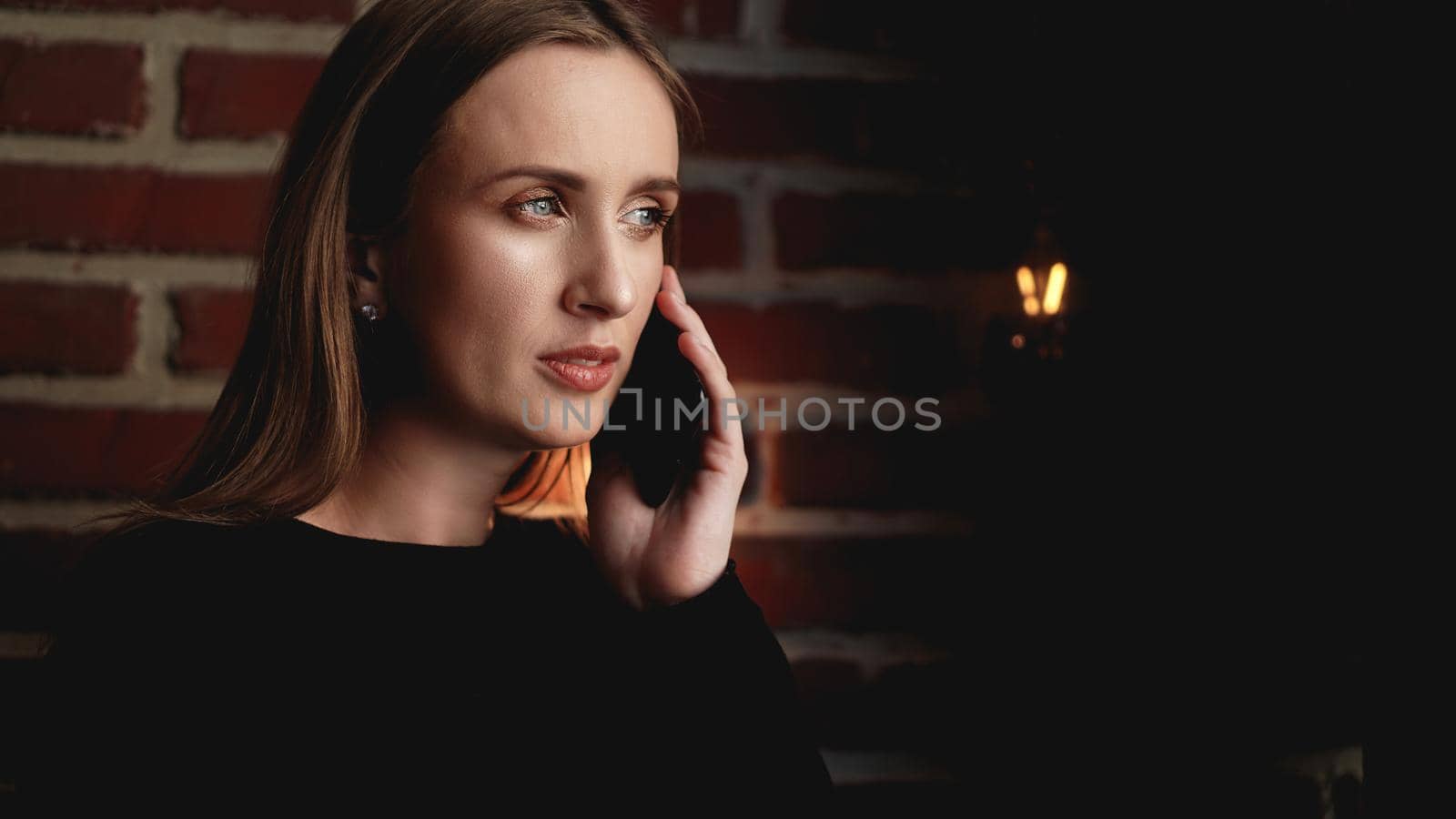 Beautiful serious young woman in black standing against brick wall and using cellphone