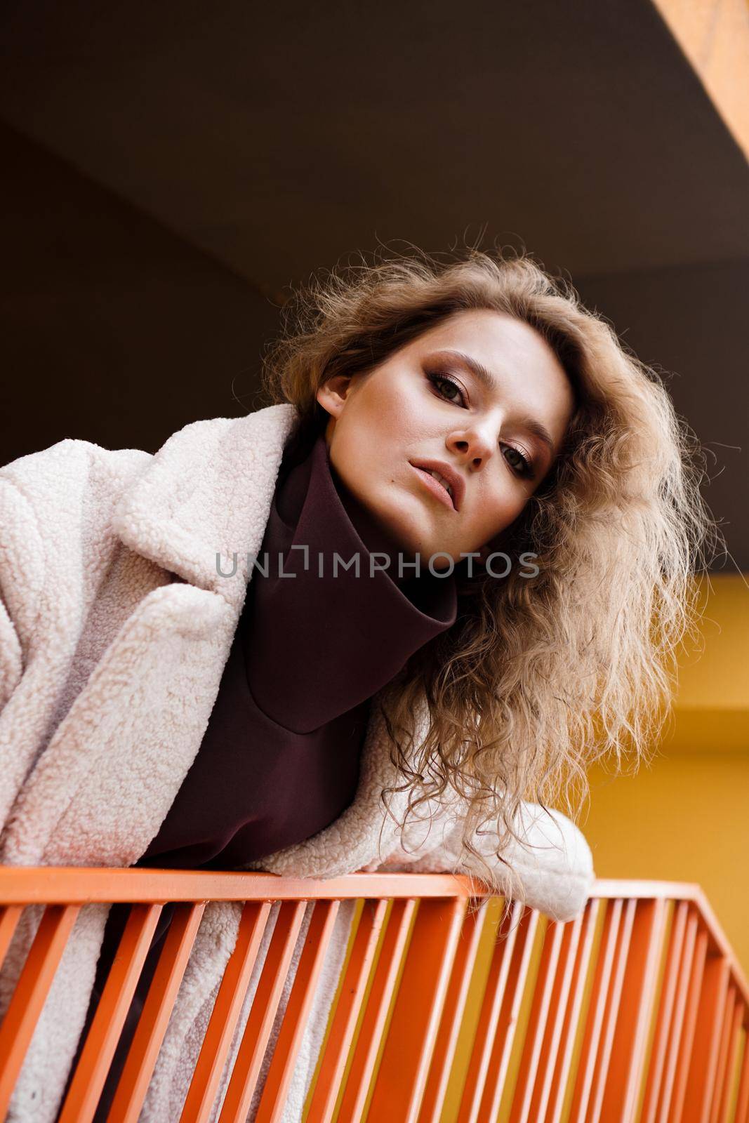 A girl with red curly hair in a white coat poses on the yellow orange parking stairs. City Style - Urban