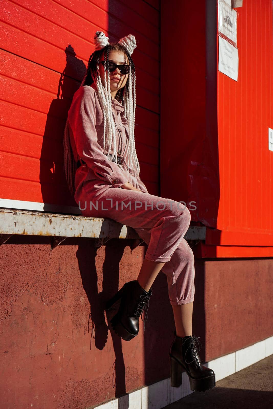 Beautiful girl over urban wall. Sunny day. Girl in sunglasses and pink jumpsuit