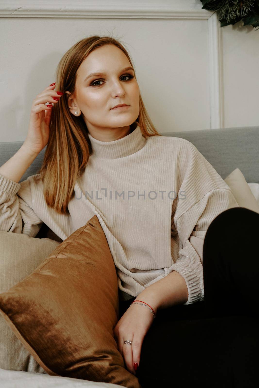 Woman with bright makeup and strict facial features dressed in beige sweater sitting on bed