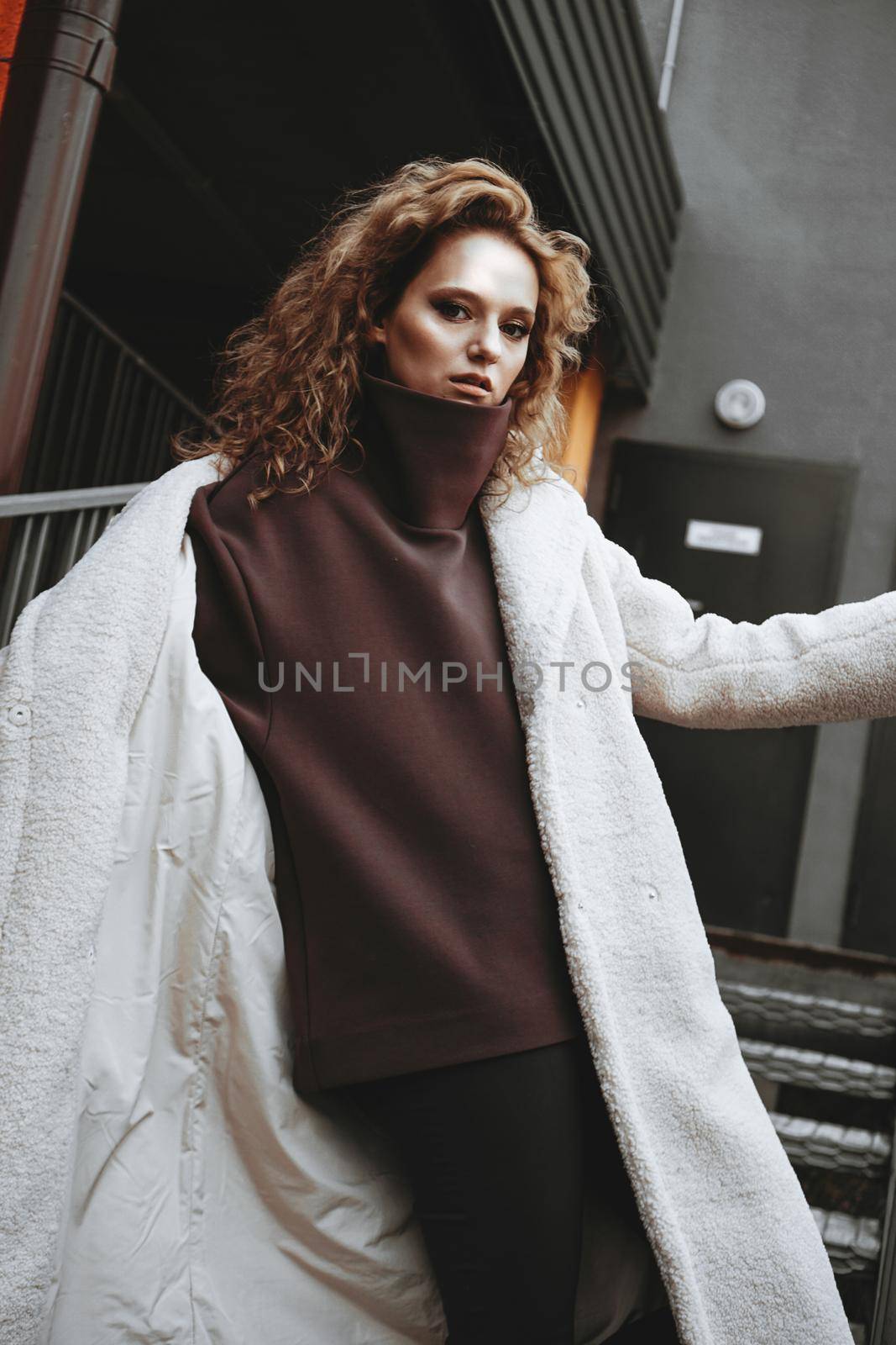 A girl with red curly hair in a white coat poses on the parking stairs. City Style - Urban