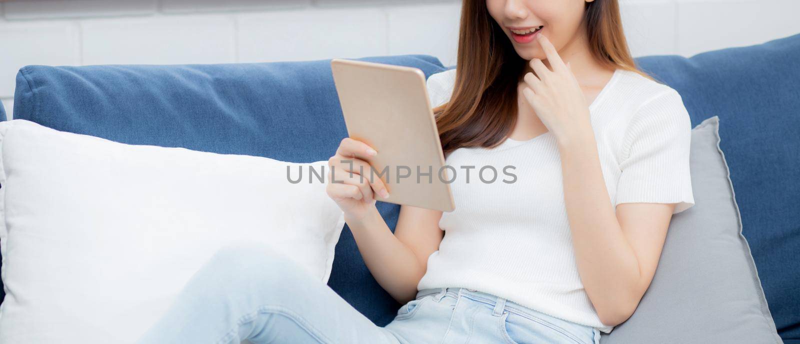 Young attractive asian woman resting using browsing tablet computer on sofa at home, happy girl sitting on couch relax reading digital gadget at house, communication and lifestyle concept.