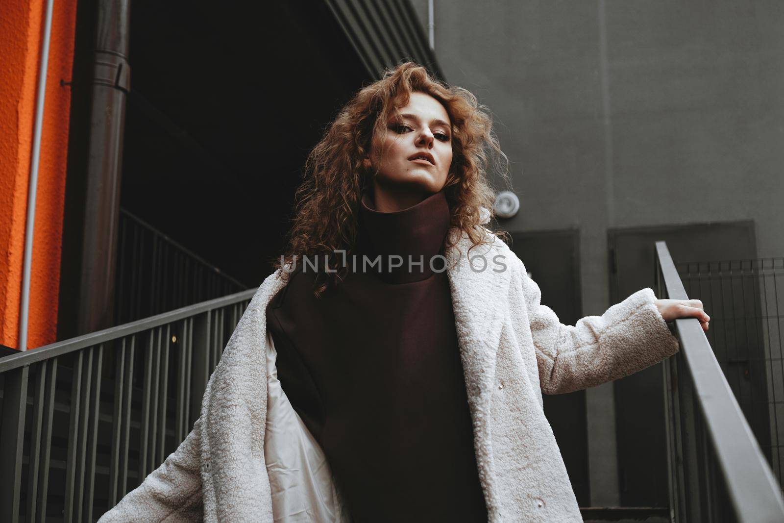 A girl with red curly hair in a white coat poses on the parking stairs by natali_brill