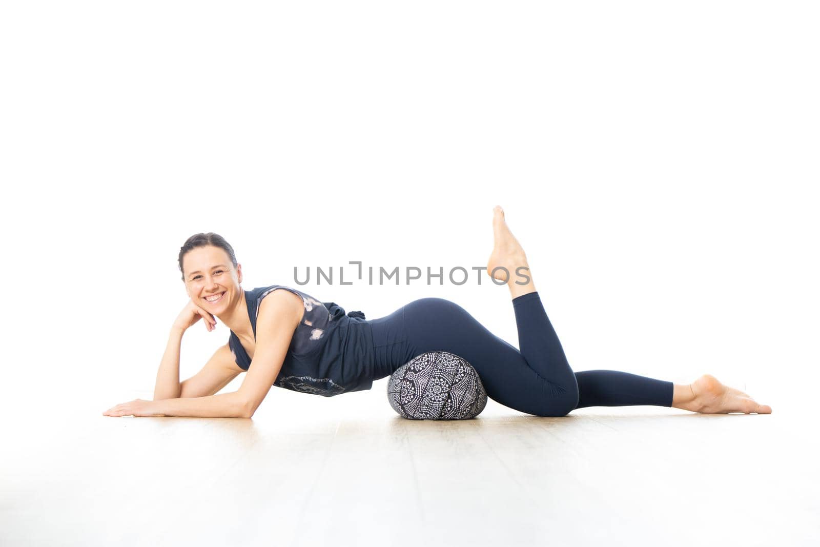 Restorative yoga with a bolster. Young sporty female yoga instructor in bright white yoga studio, lying on bolster cushion, stretching, smilling, showing love and passion for restorative yoga.