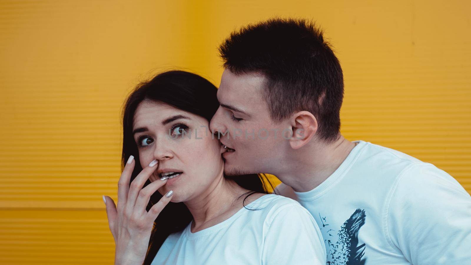 Funny young man bites his girlfriends ear and embracing her. Couple having fun - yellow background