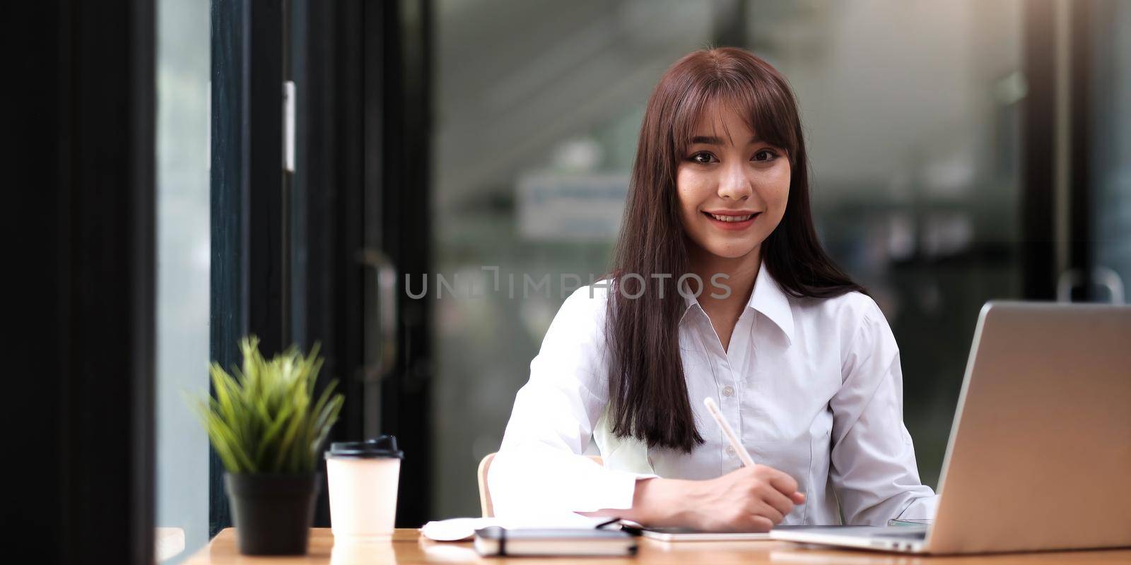 Portrait of a pretty young woman studying while sitting at the table with laptop computer and notebook at home by wichayada