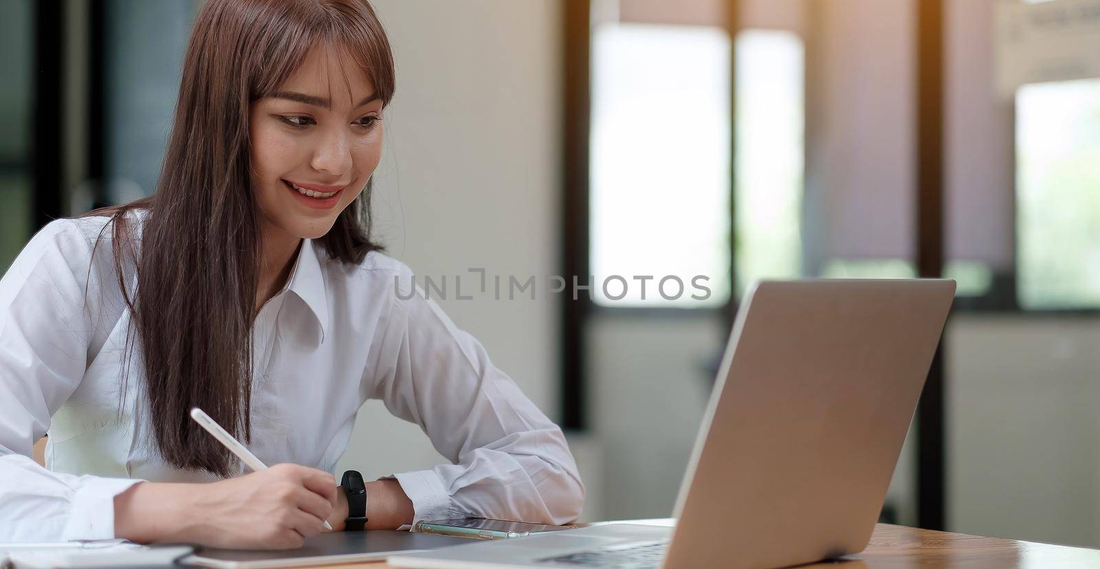A young, cheerful girl in a white shirt smiles, smiling toothily writing down notes holding training for students to be executives at laptop desktop table by wichayada