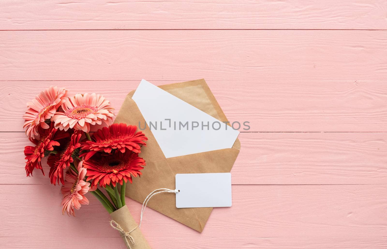 Happy birthday. Red gerbera daisy flowers, envelope and blank label tag on pink wooden table