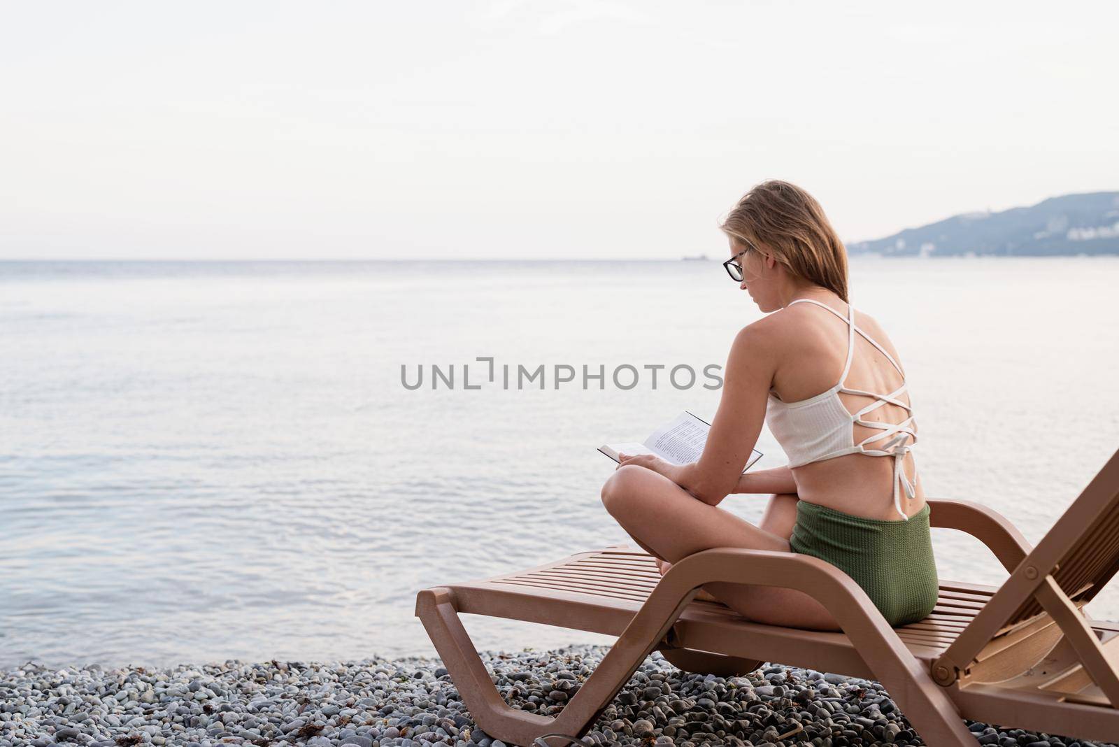 Summer vacation on the beach. The beautiful young woman in swimming suit sitting on the sun lounger reading a book and relaxing, blue hour