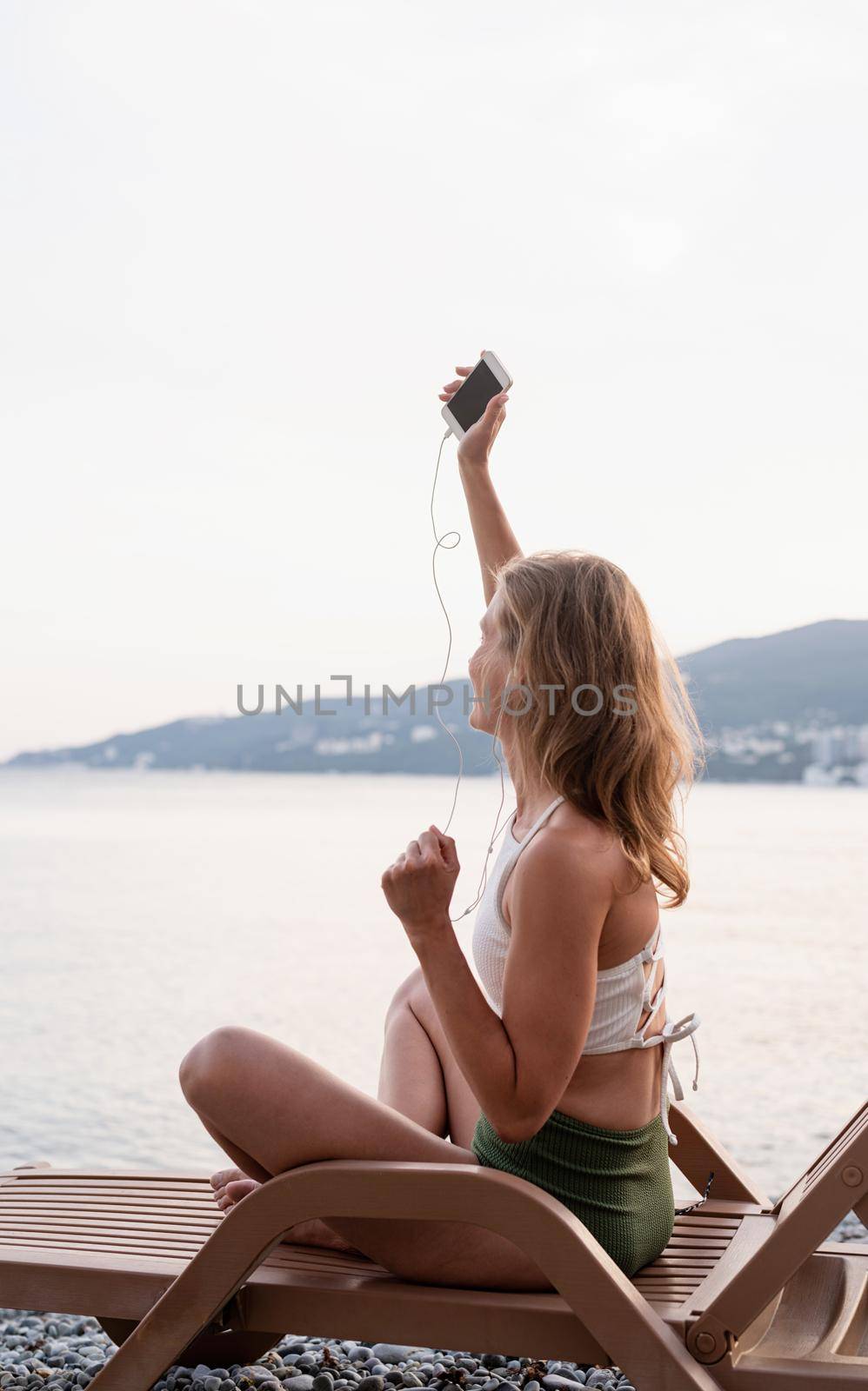 Summer vacation on the beach. The beautiful young woman in swimsuit sitting on the sun lounger listening to the music using mobile device and dancing, blue hour
