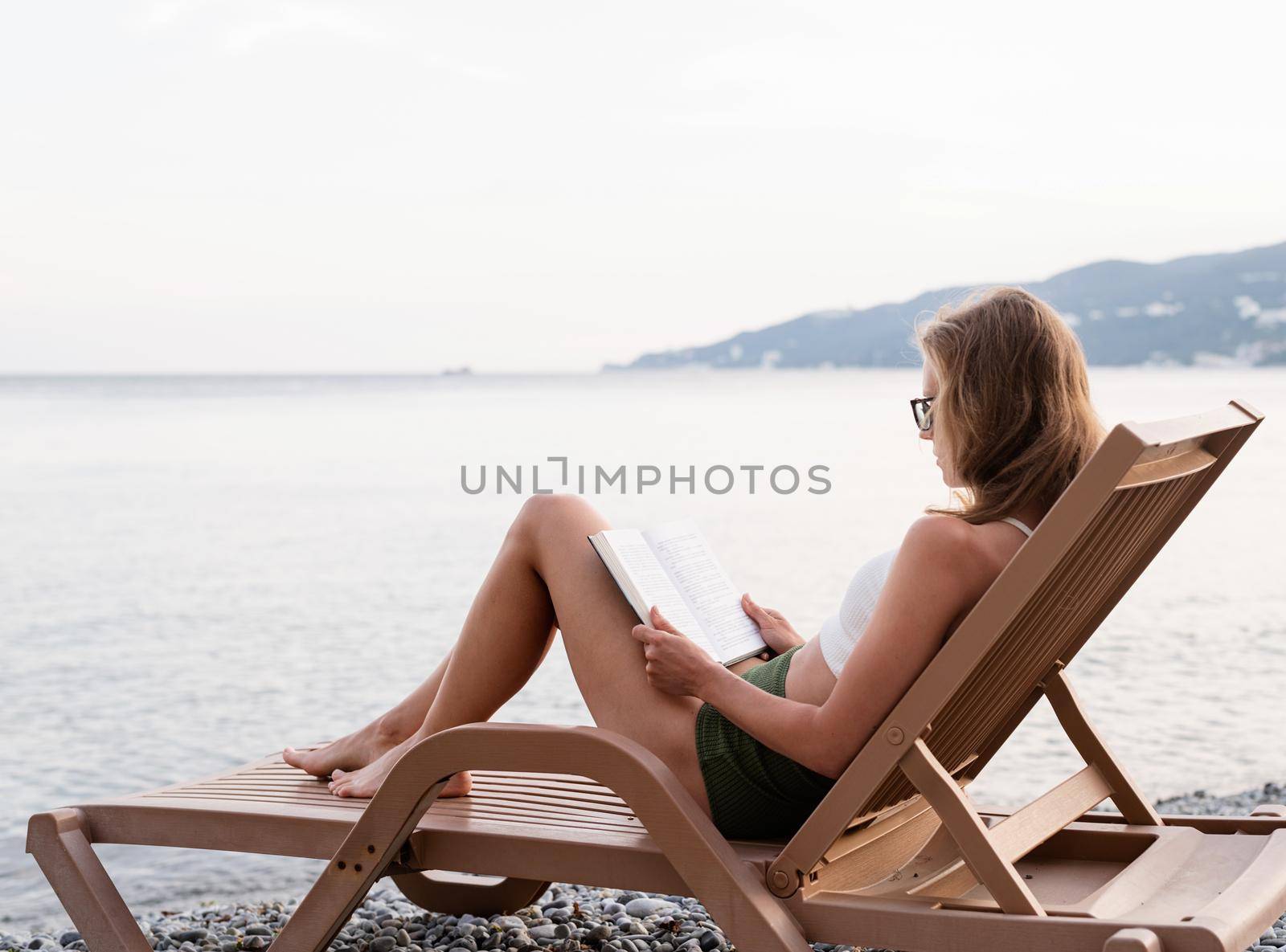Summer vacation on the beach. The beautiful young woman in swimming suit sitting on the sun lounger reading a book and relaxing