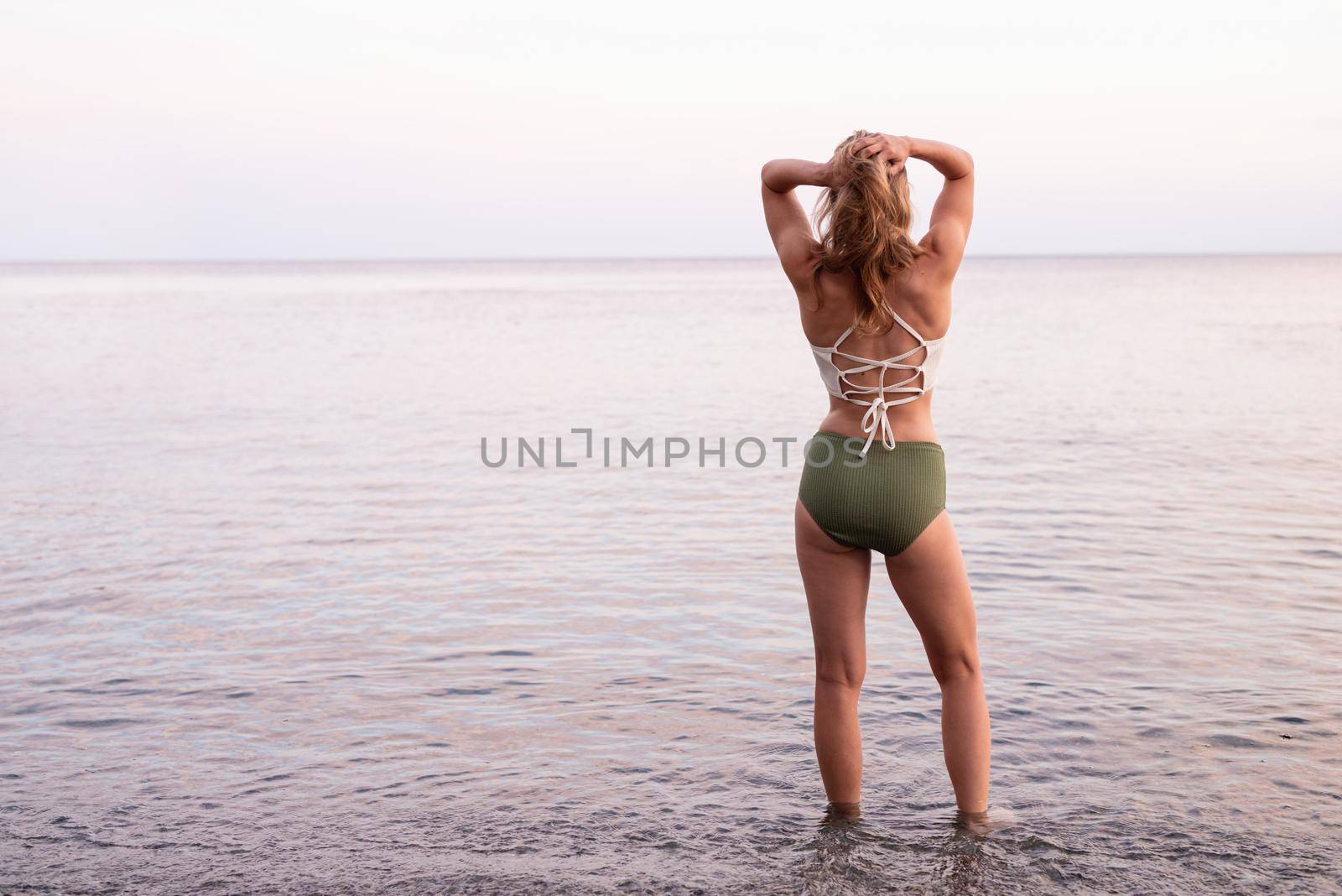 Summer vacation. Rear view of a young woman standing on stony beach looking at the sea, hands on head