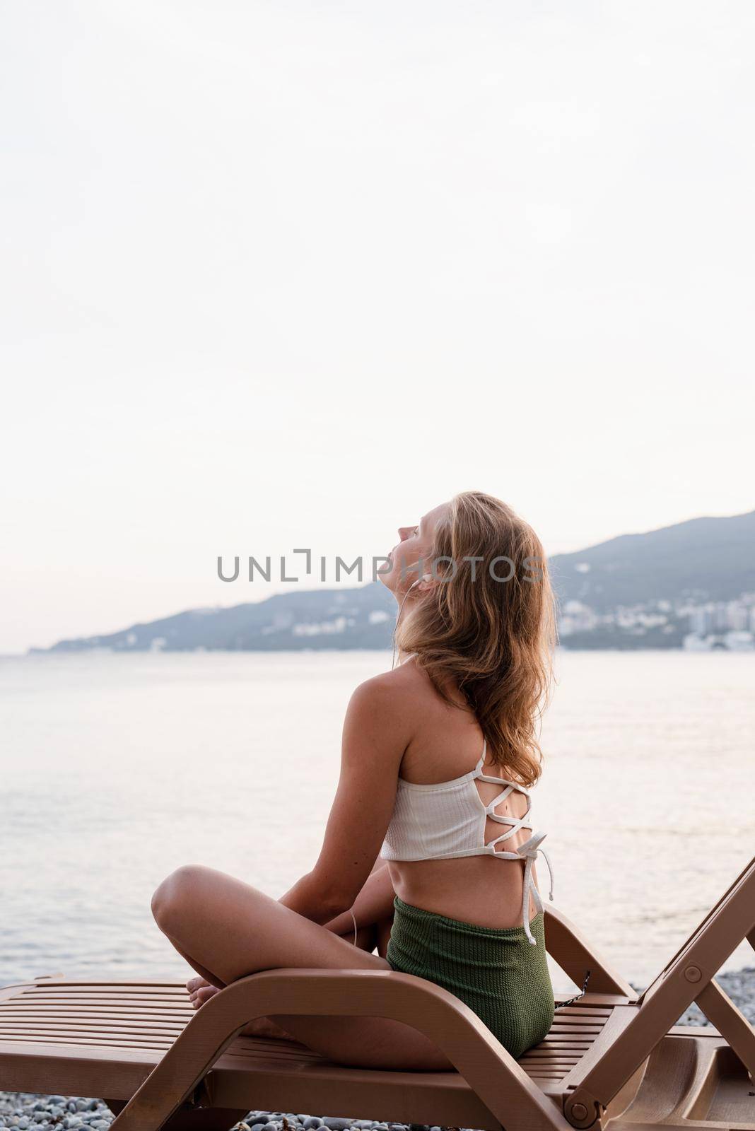 Summer vacation on the beach. The beautiful young woman in swimsuit sitting on the sun lounger listening to the music using mobile device and dancing, blue hour
