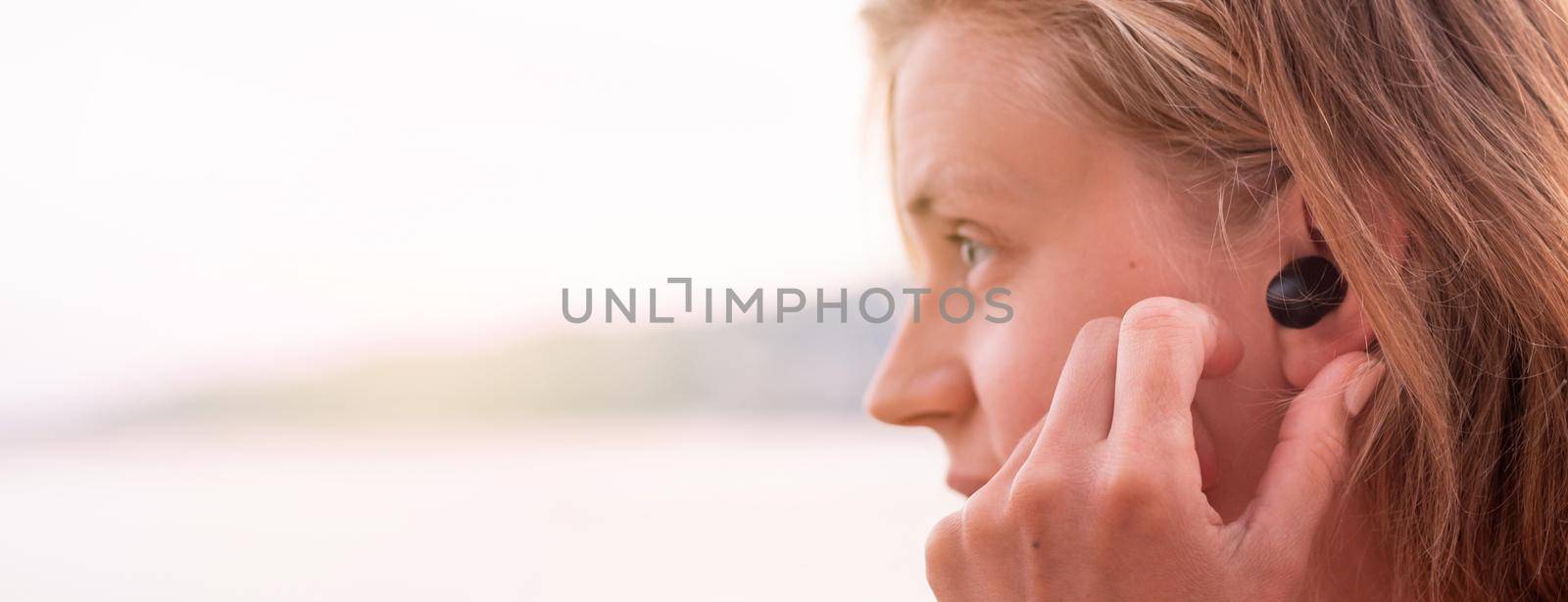 Technology concept. Woman using wireless earbuds, seaside on the background, banner