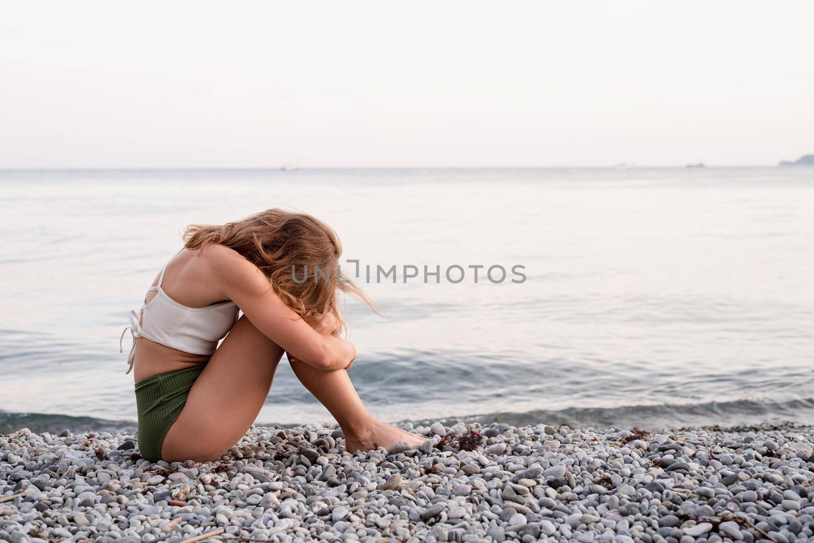 Mental health concept. Rear view of a depressed young woman in swimsuit sitting on the beach looking away
