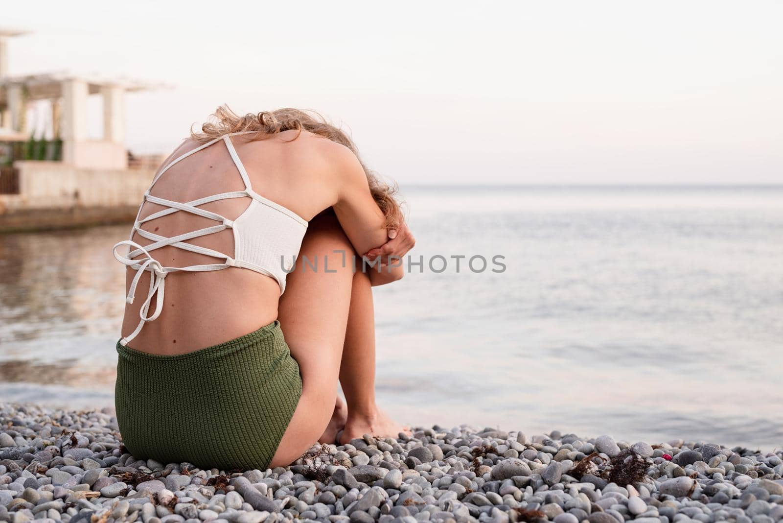Mental health concept. Rear view of a depressed young woman in swimsuit sitting on the beach, arms around legs