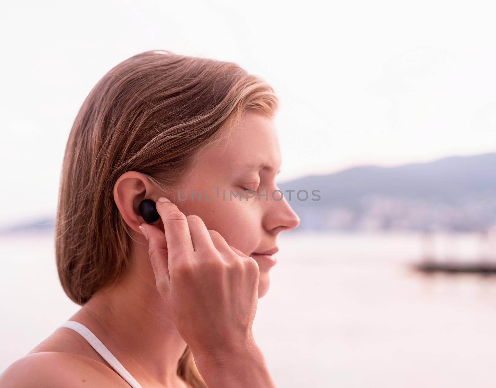 Technology concept. Woman using wireless earbuds, seaside on the background
