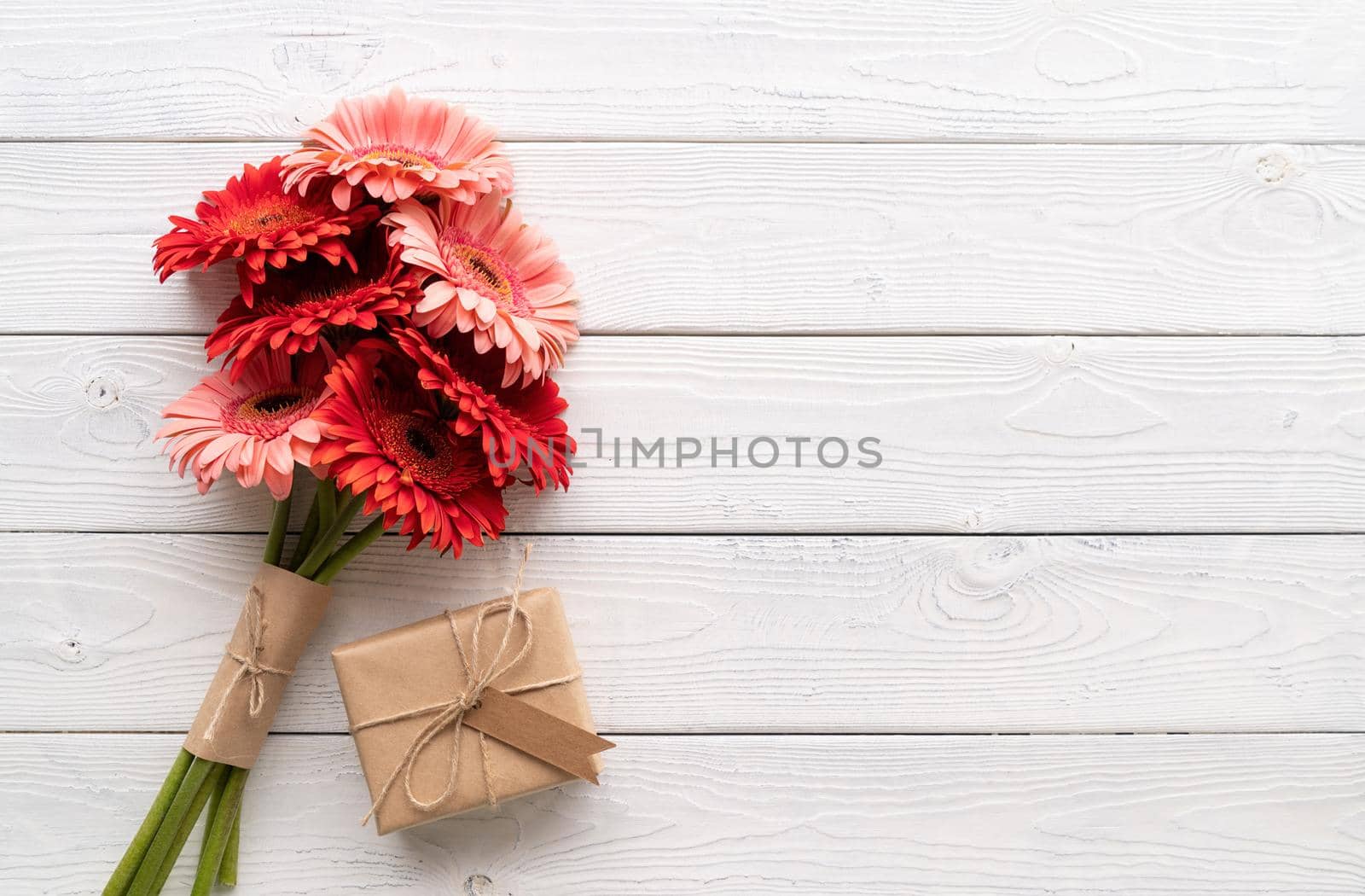 Happy birthday. Red gerbera daisy flowers and craft gift box with label tag on white wooden table, flat lay
