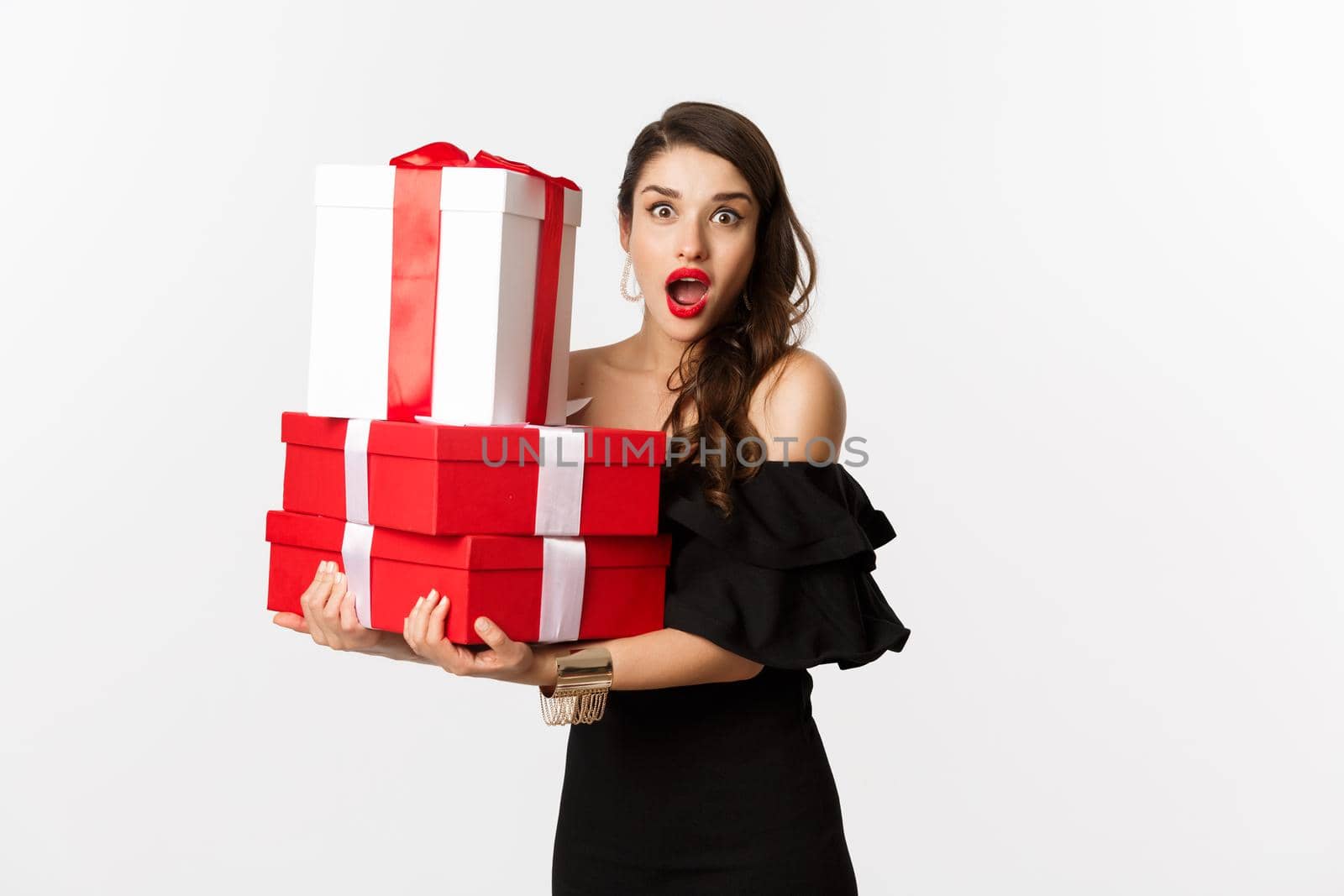 Celebration and christmas holidays concept. Beautiful woman in black dress holding gifts and looking surprised, standing over white background by Benzoix