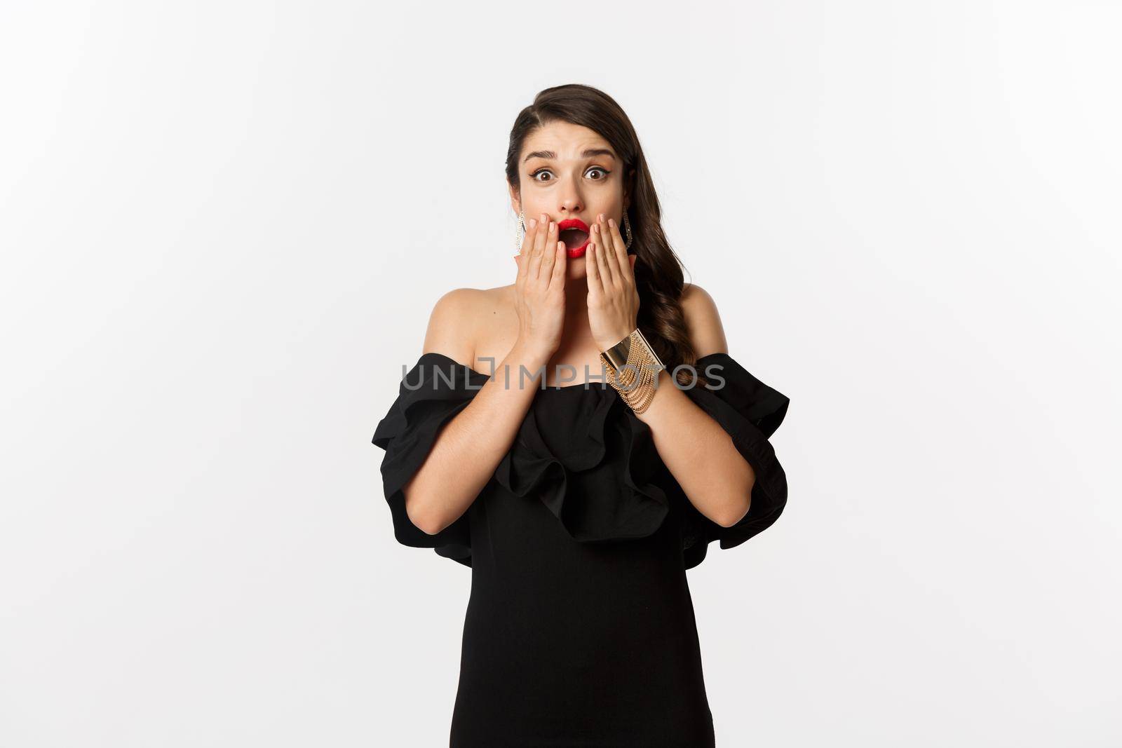 Fashion and beauty. Image of attractive female model in black dress reacting to announcement, looking amazed at camera, standing surprised over white background.