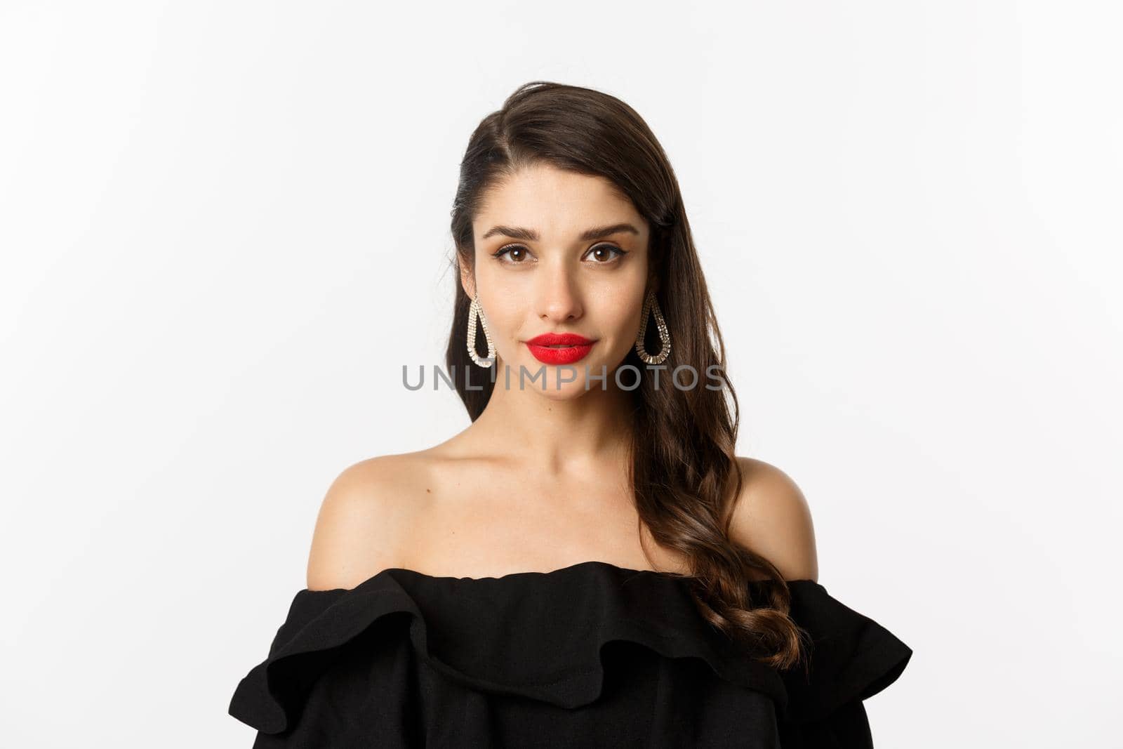 Fashion and beauty concept. Close-up of elegant brunette woman with earrings, wearing black dress and red lipstick, looking sensual at camera, standing over white background by Benzoix