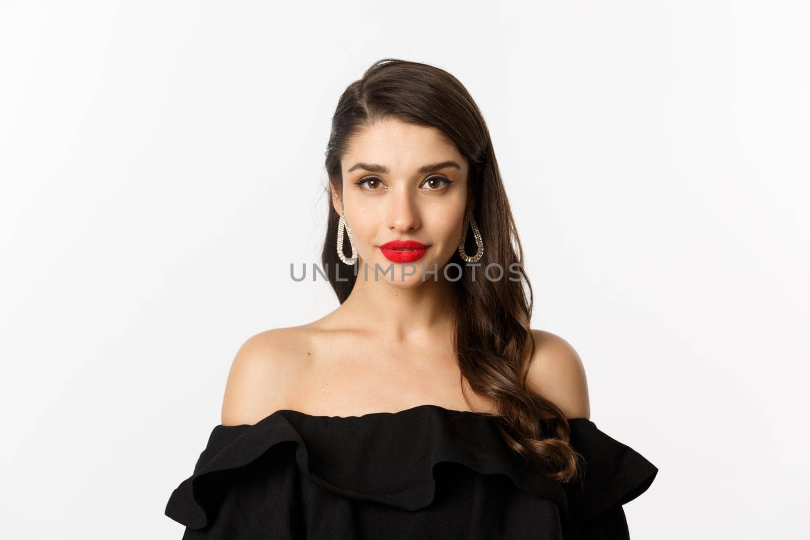 Fashion and beauty concept. Close-up of elegant brunette woman with earrings, wearing black dress and red lipstick, looking sensual at camera, standing over white background.