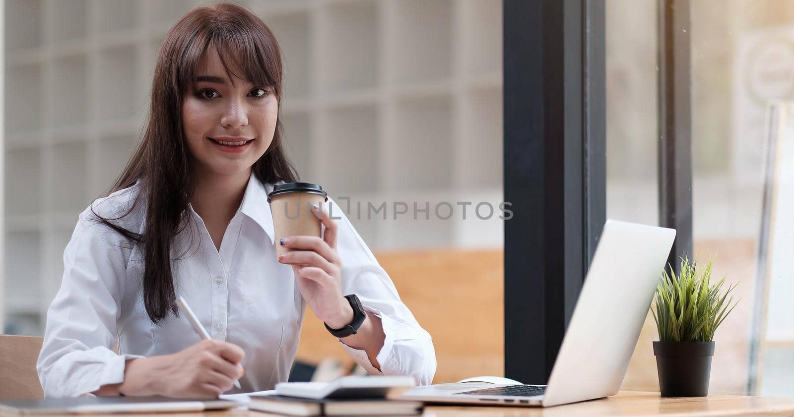 Woman having a tea, sitting on a floor in an office, writing down notes, opened laptop in front of her. Home office concept.