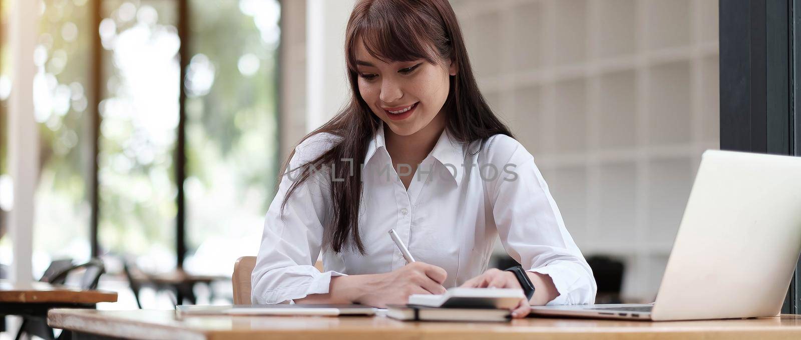 A young, cheerful girl in a white shirt smiles, smiling toothily writing down notes holding training for students to be executives at laptop desktop table by wichayada