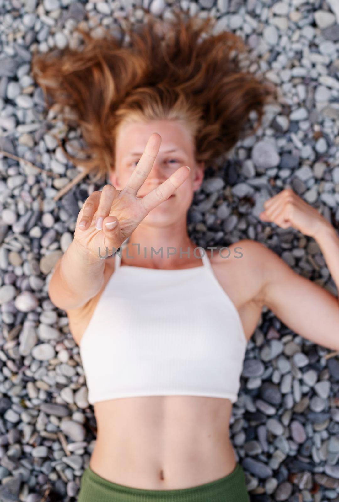 Top view of young smiling woman lying on stone beach, covering face with hands by Desperada