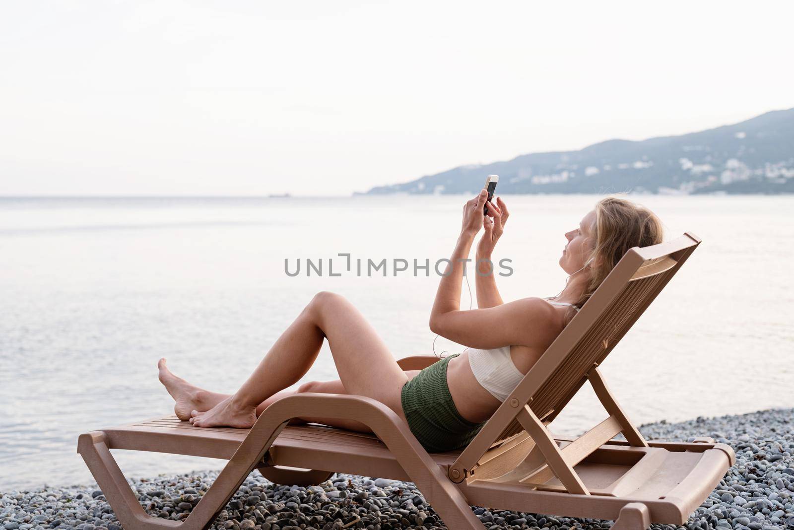 Summer vacation on the beach. The beautiful young woman in swimsuit lying on the sun lounger listening to the music using mobile device, blue hour