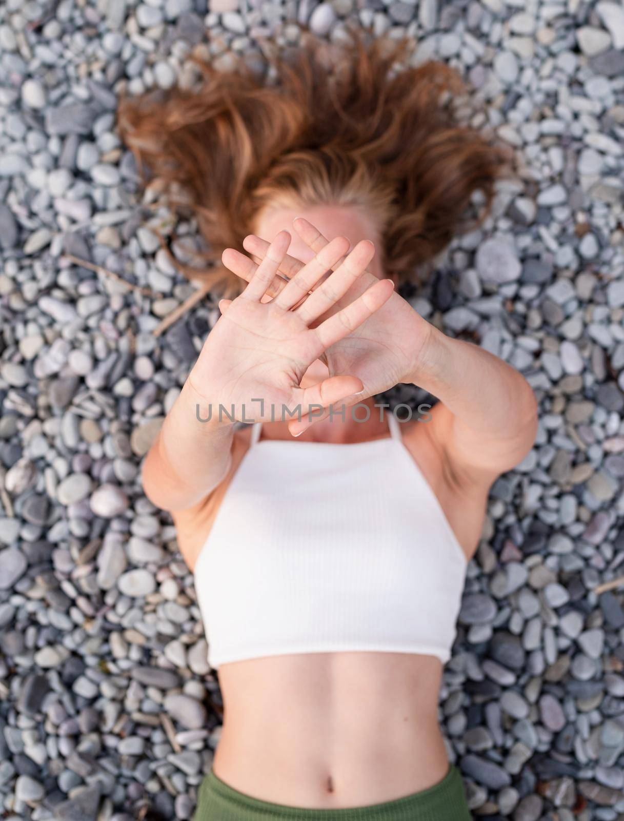 Top view of young smiling woman lying on stone beach, covering face with hands by Desperada