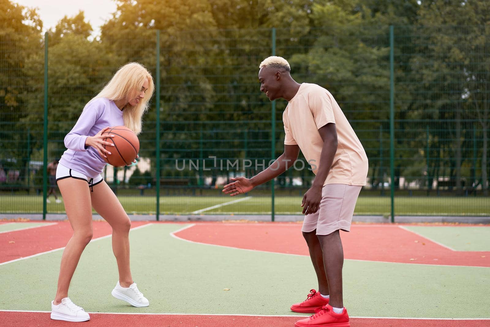 Active multiethnic couple playing basketball on outdoor sport court, black guy teaching his cute blonde girlfriend to play streetball Multinational relationship