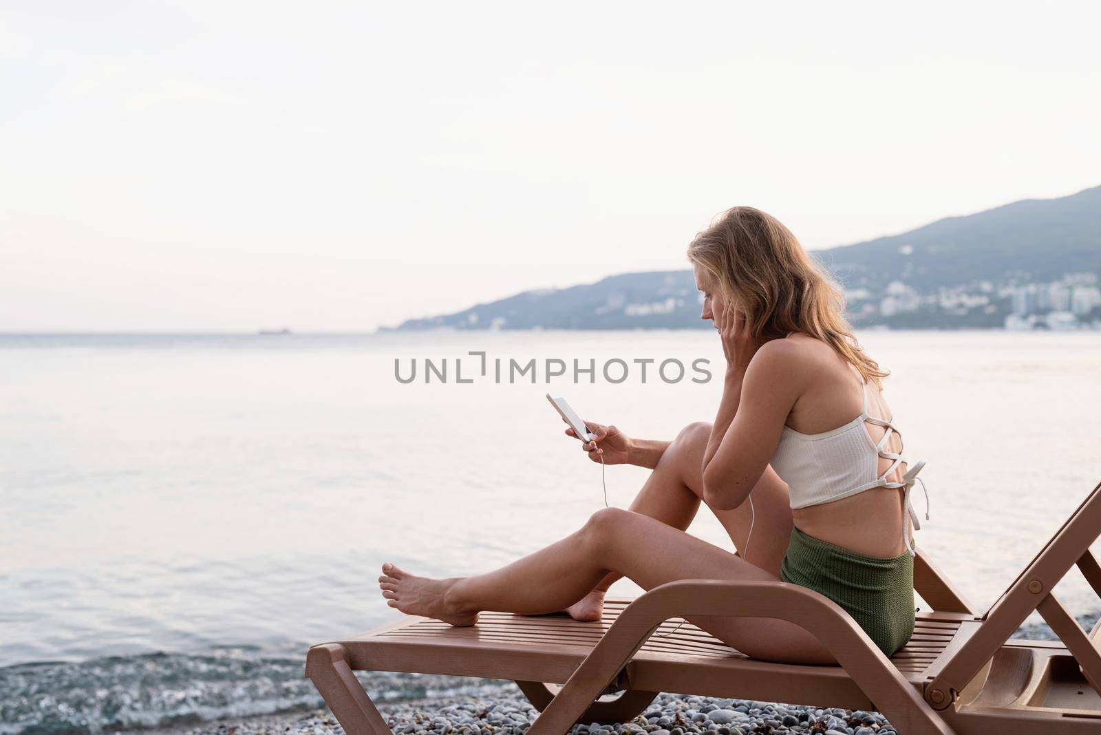 Summer vacation on the beach. The beautiful young woman in swimsuit sitting on the sun lounger listening to the music using mobile device, blue hour