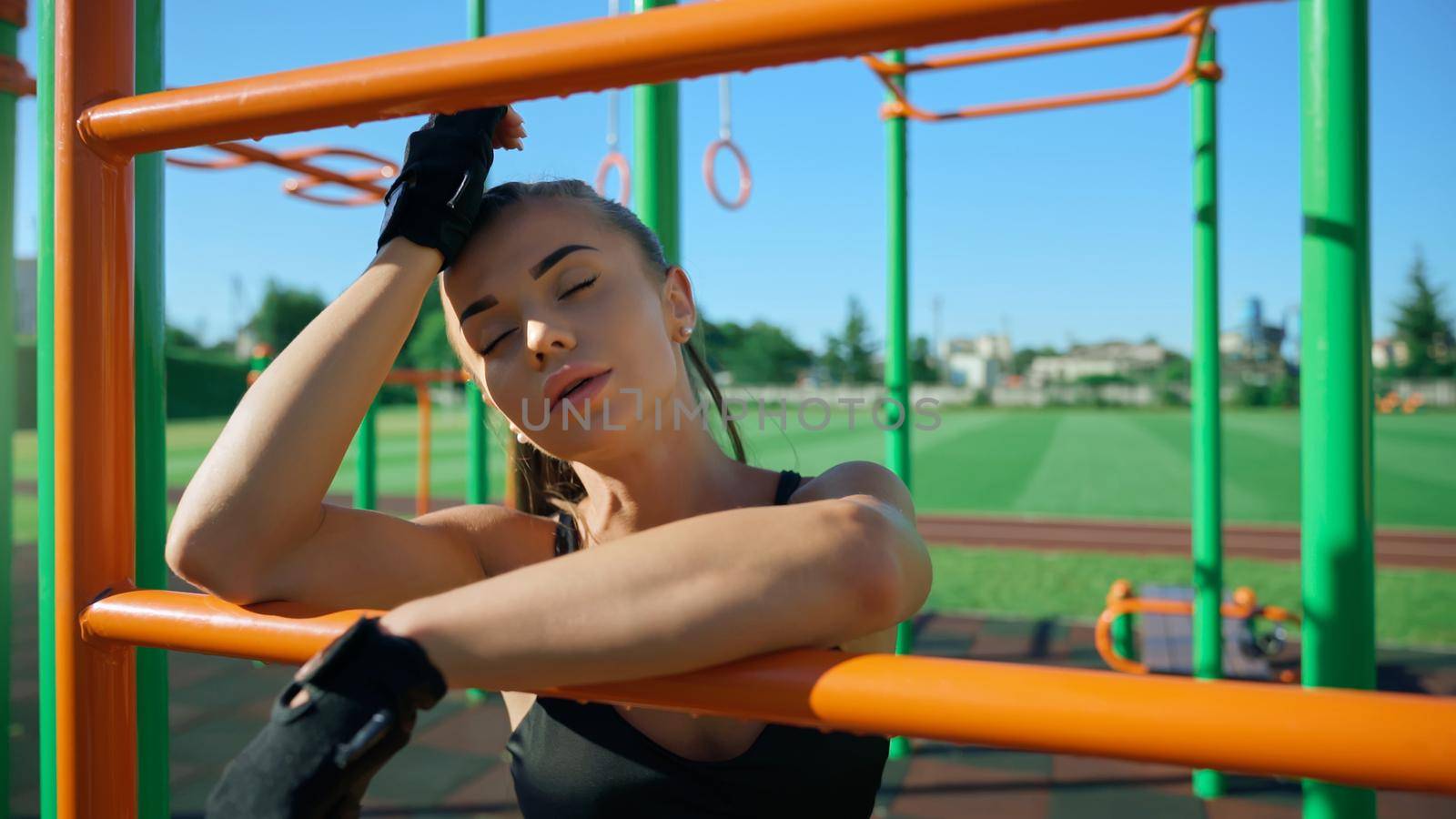 Young sexy woman with eyes closed posing near bars at sports ground in summer sunny day, touching face. Sportswoman with perfect makeup wearing fashionable outfit outdoors. Workout concept.