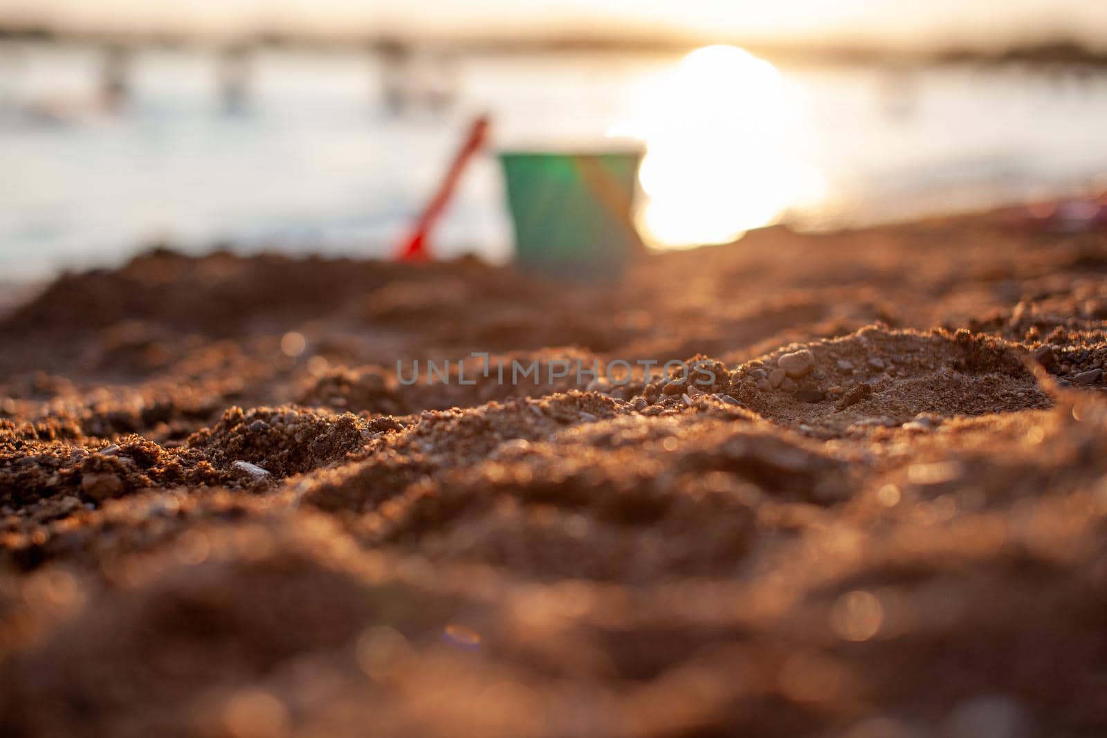 Children's toys for playing on the sand. Plastic bucket and rake on the beach at sunset. The concept of summer, family holidays and vacations. The bucket is out of focus. Focus on the sand