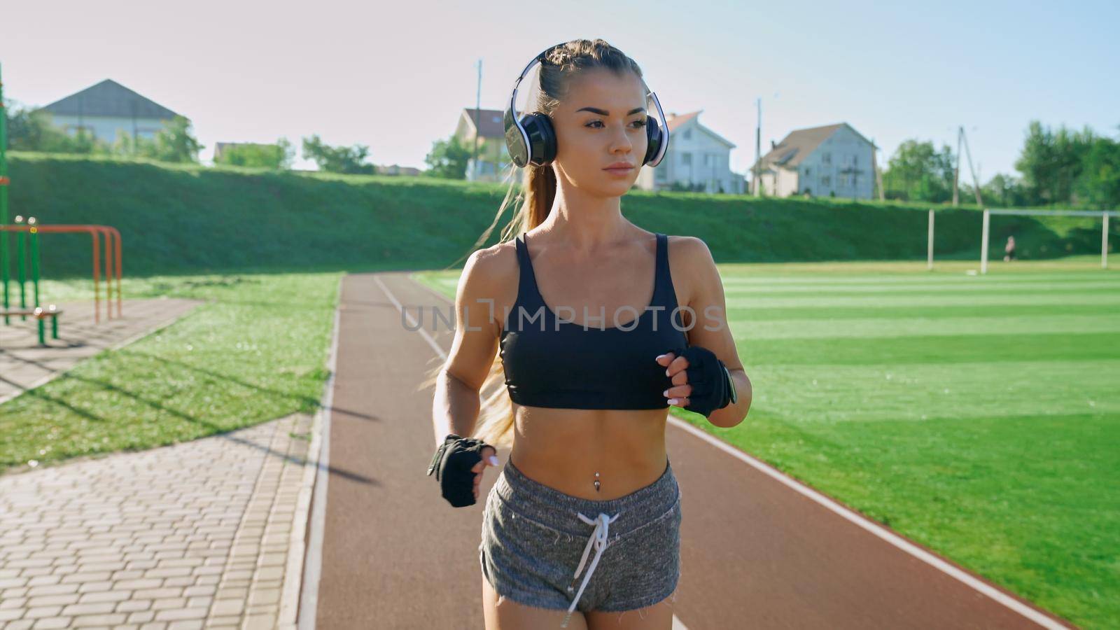 Front view of young athlete woman wearing big wireless headphones and black gloves running outdoors. Stunning girl with pony tail practicing jogging at stadium in sunny summer morning.