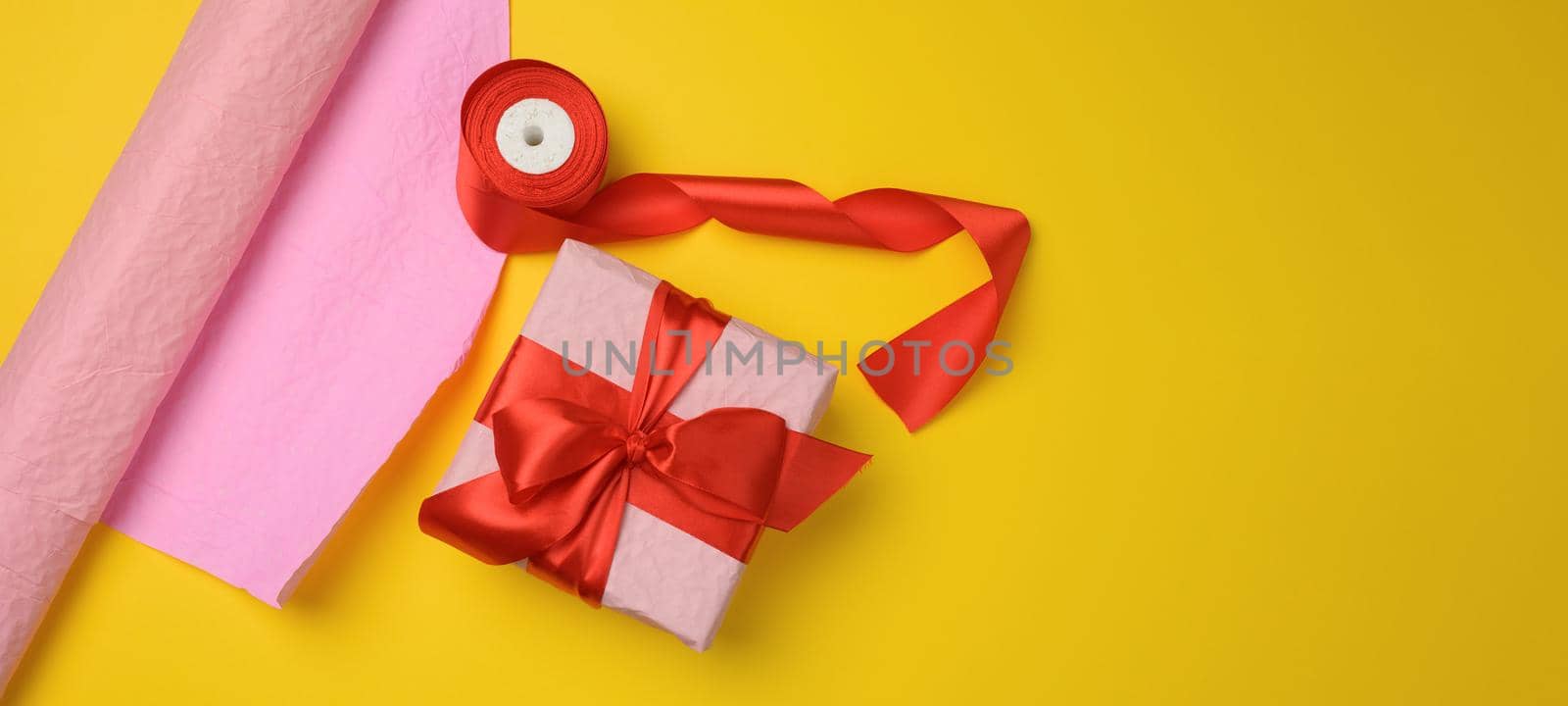 pink gift box wrapped in red silk ribbon on yellow background, copy space
