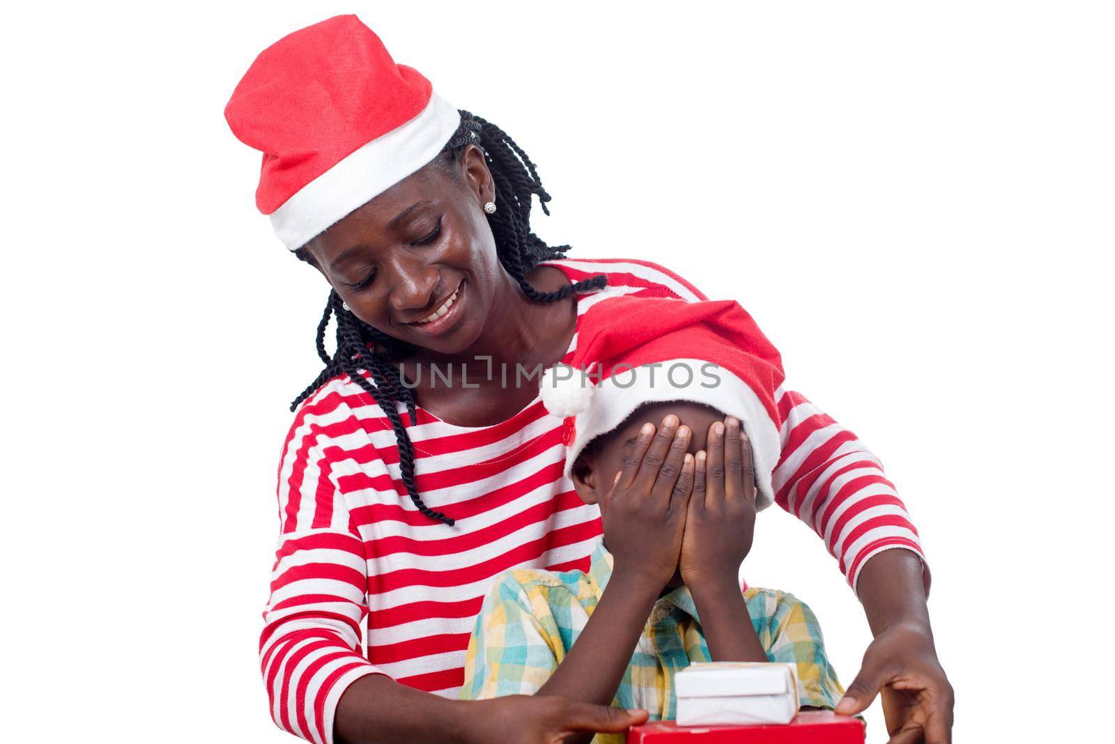 smiling young woman in Santa Claus hat holds a gift in front of her child in Santa Claus hat and eyes closed by her hands.