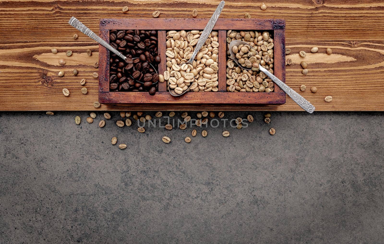 Green and brown unroasted and dark roasted coffee beans in wooden box with spoons setup on dark concrete background.