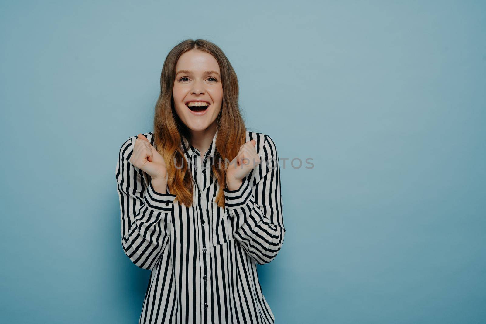 Overjoyed young woman wearing casual clothes feeling very happy and excited, doing winner gesture with clenched, screaming while celebrating success while posing isolated on blue background