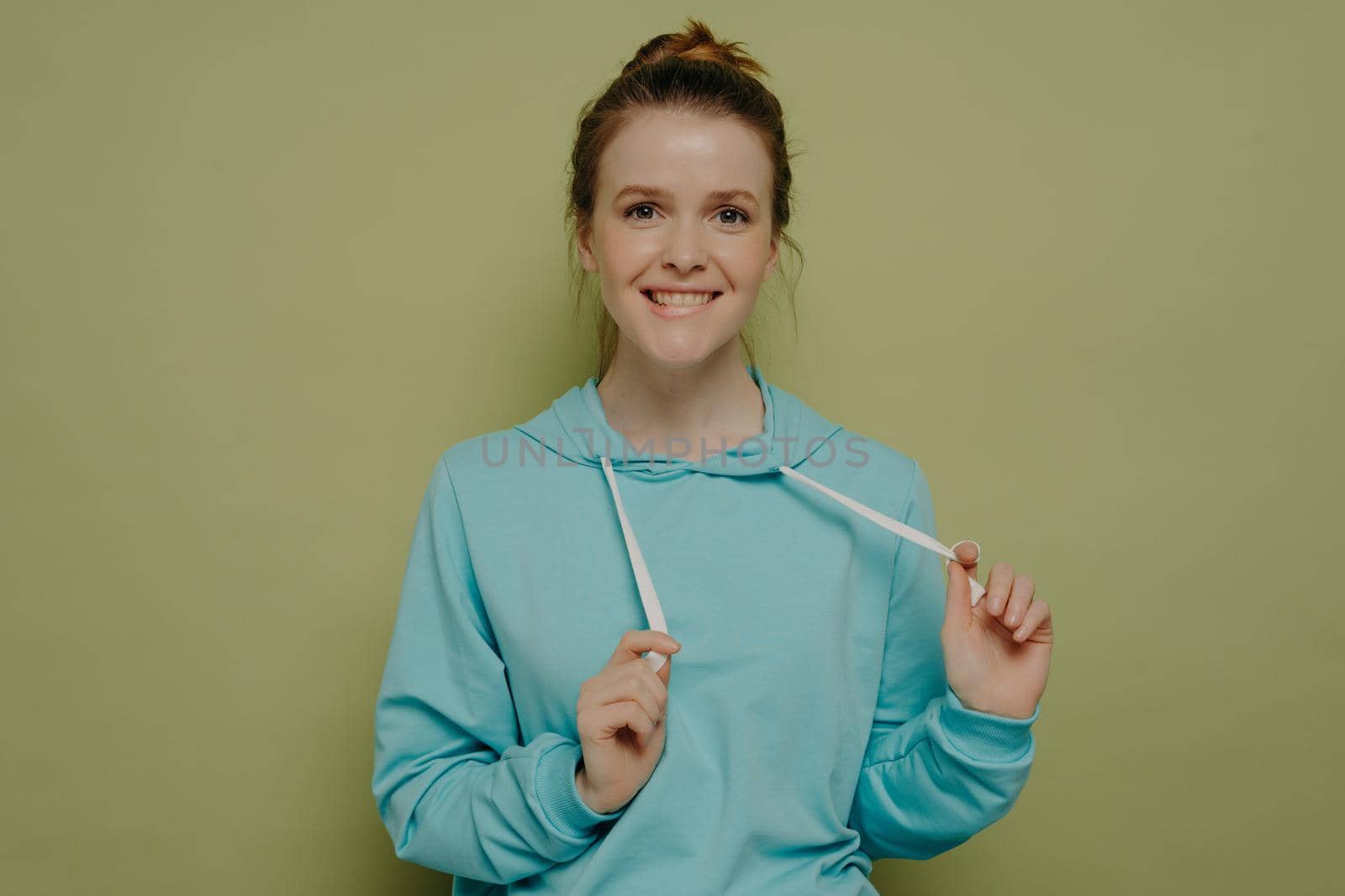 Portrait of cute flirty teenage girl biting lower lip playing with hoodie strings, standing in front camera on color background in studio, female feeling playful and carefree